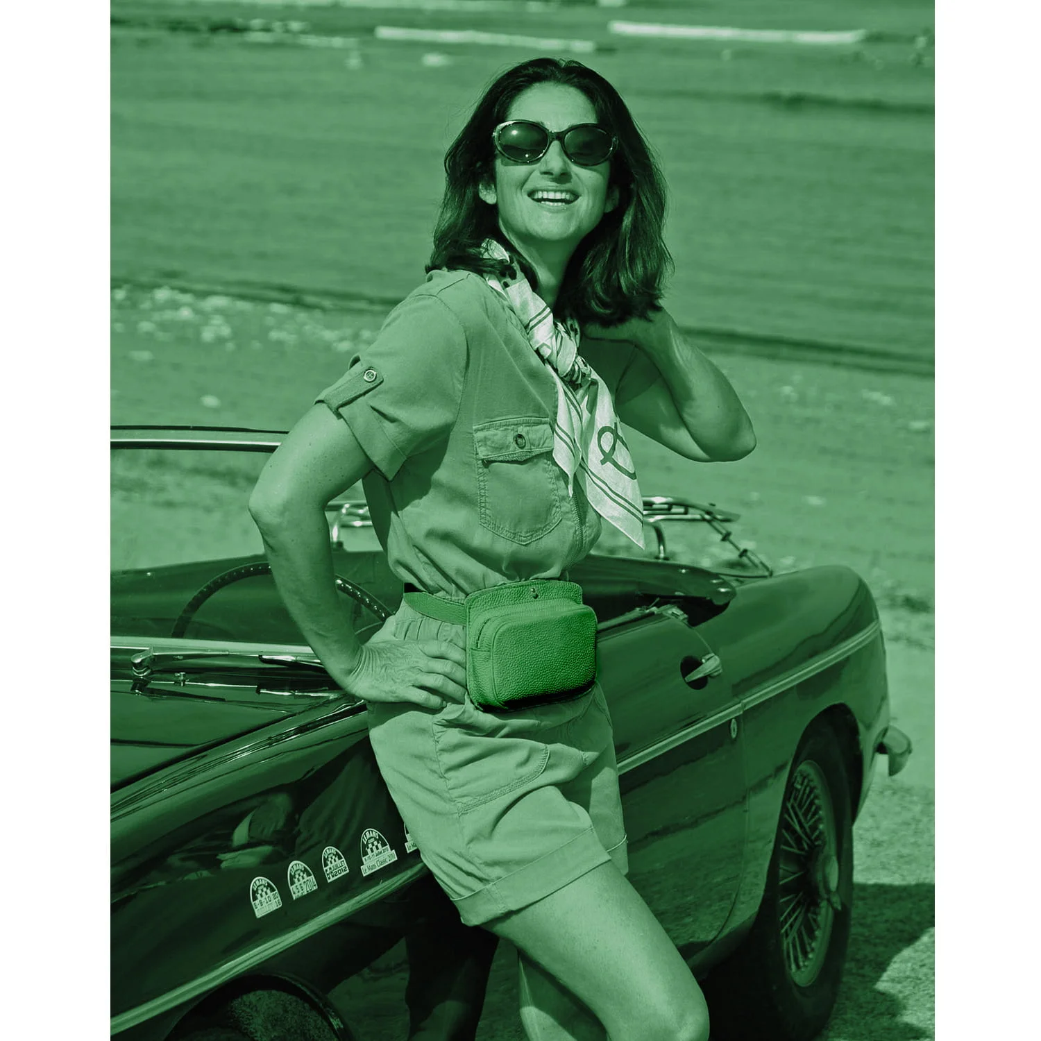 Une personne portant des lunettes de soleil pose à côté d'une décapotable classique sur la plage. Elle porte un foulard, une combinaison et un sac banane, et sourit à la caméra.