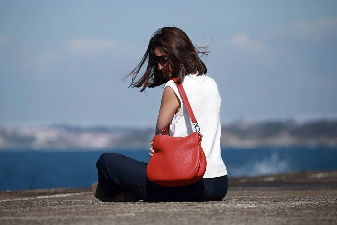 Une femme portant des lunettes de soleil est assise au bord de l'eau, le dos tourné, tenant un sac rouge sur son épaule.