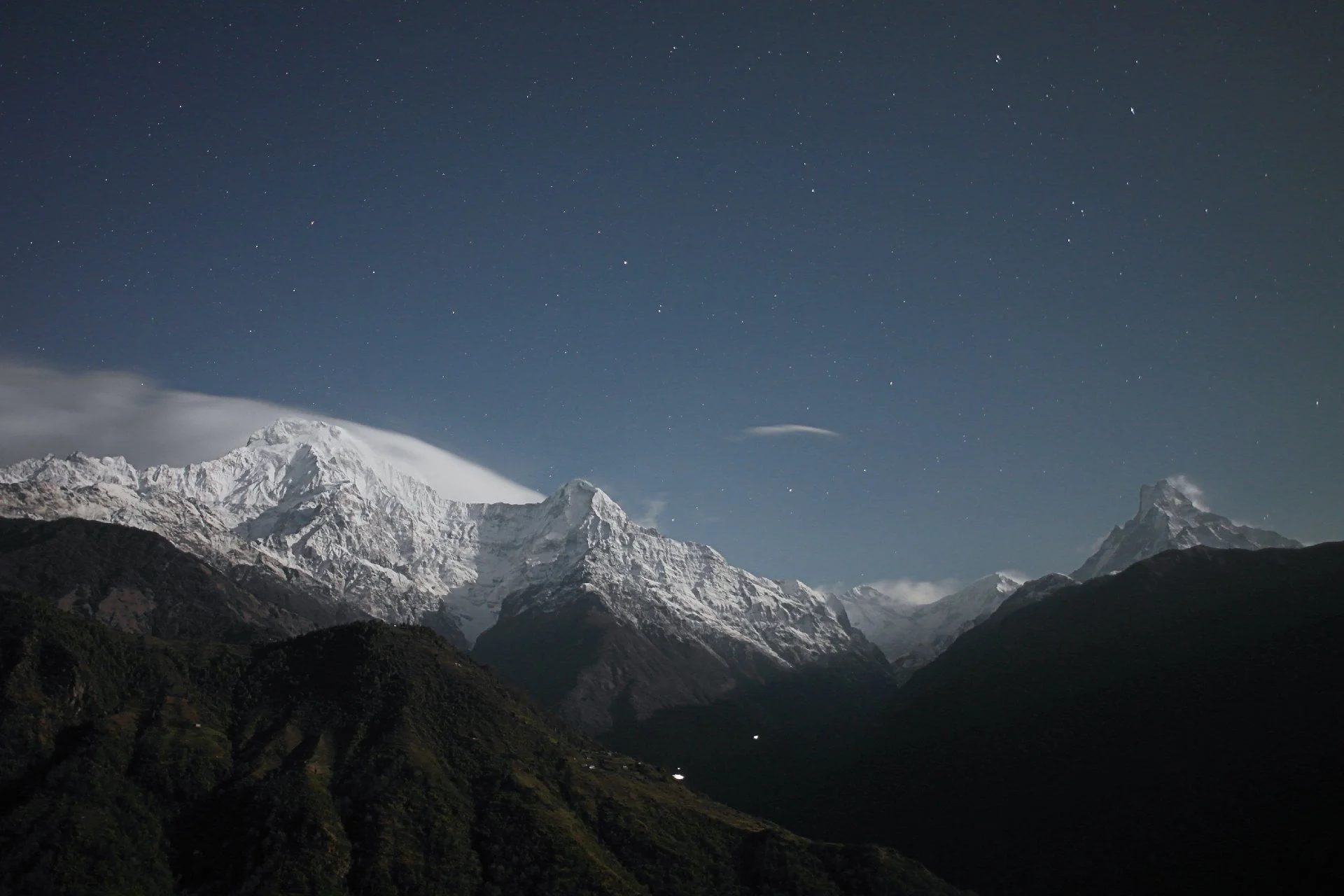 Montagnes enneigées sous un ciel étoilé avec une petite habitation visible à la base.