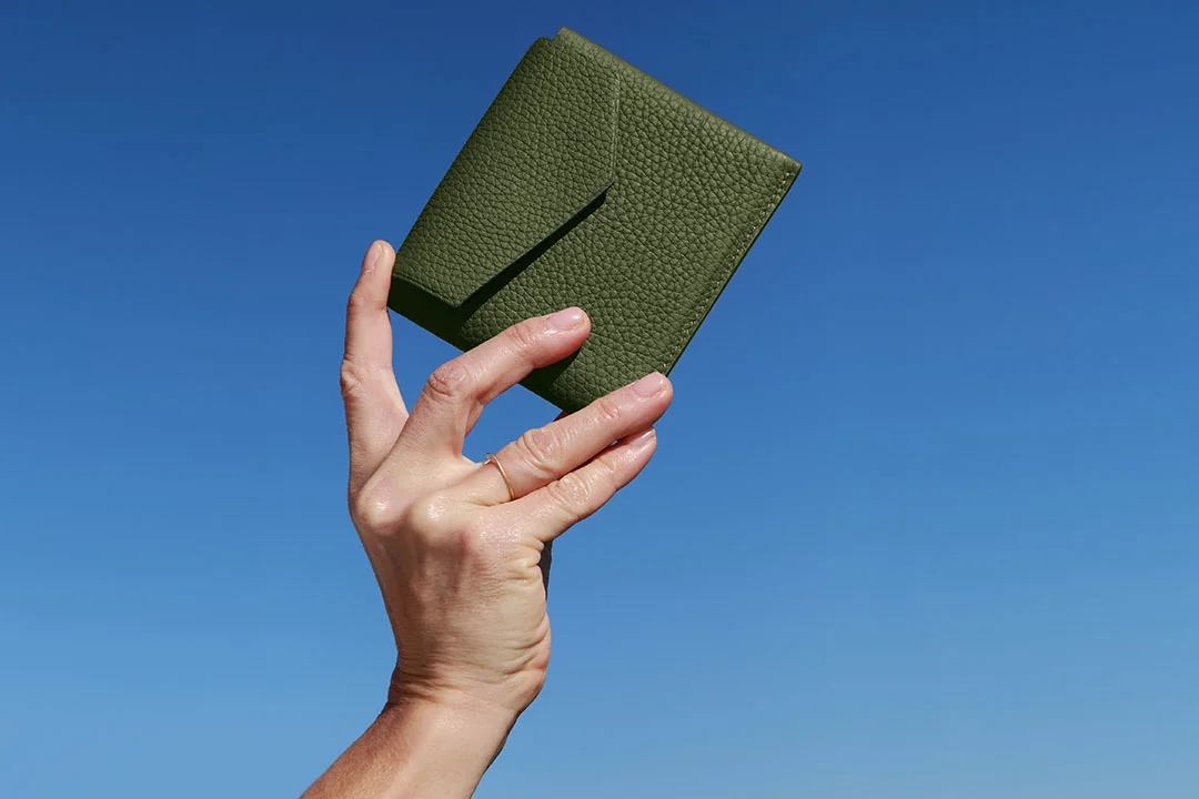 Une main tenant un portefeuille en cuir vert sur un fond de ciel bleu clair.