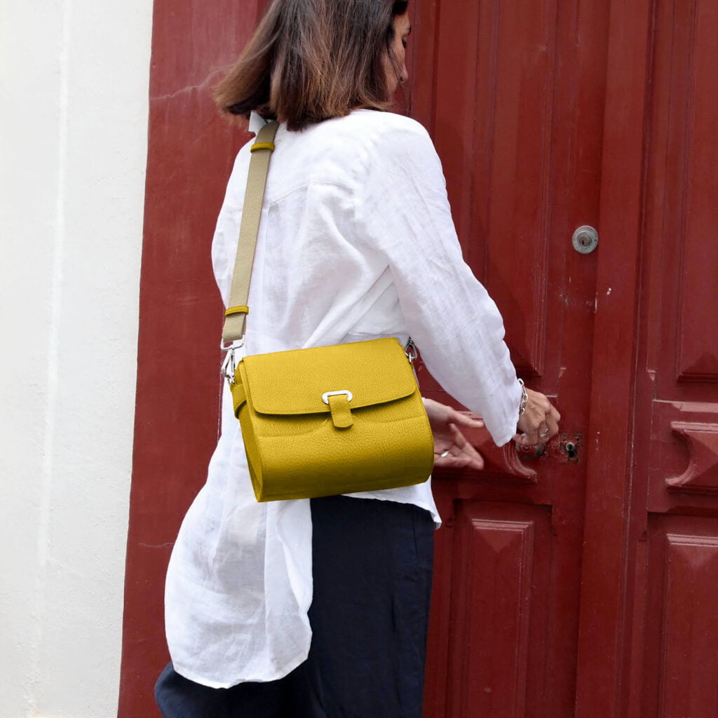 Une personne portant une chemise blanche et un pantalon foncé se tient devant une porte rouge, portant un sac à bandoulière jaune.