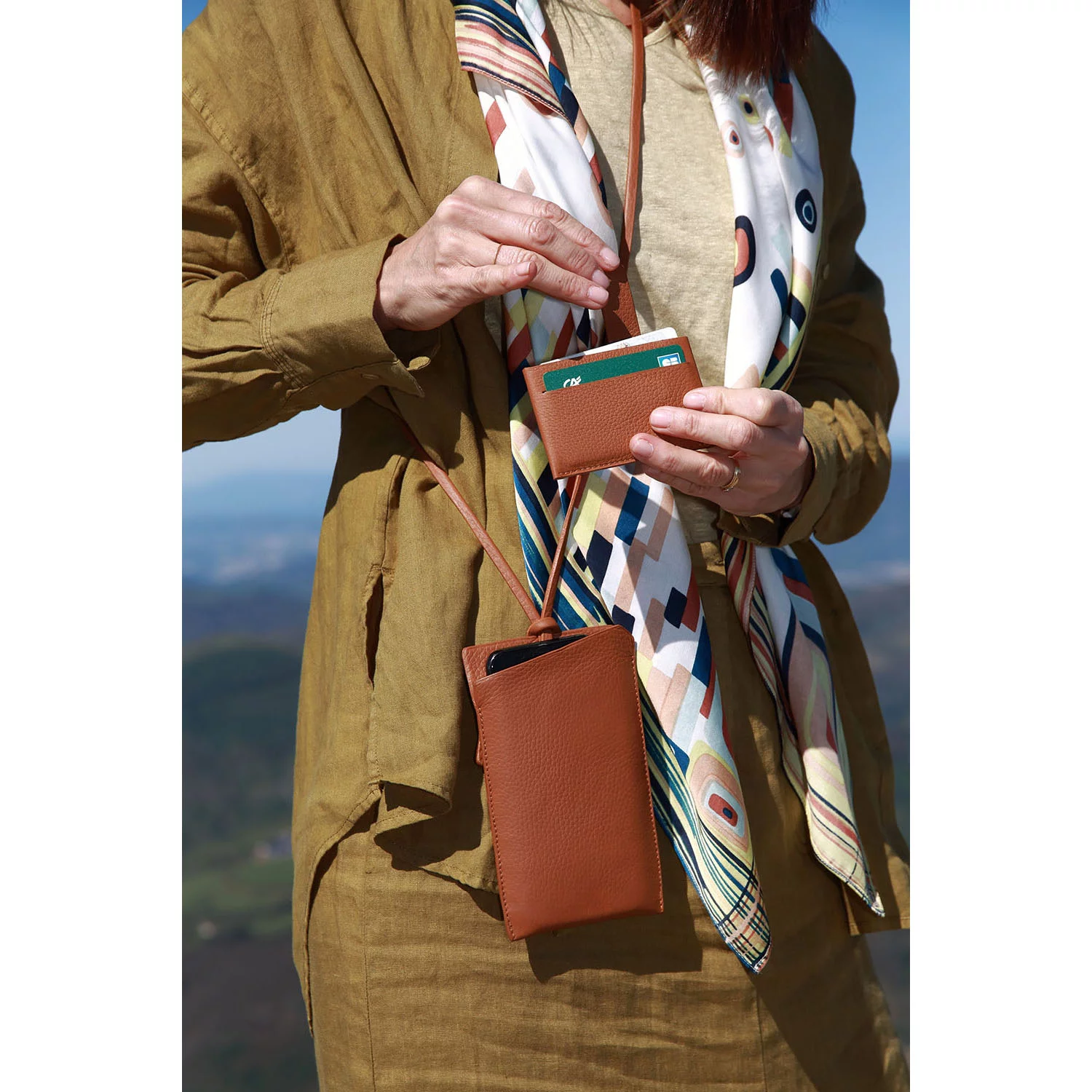 Personne tenant un portefeuille marron et une pochette de téléphone, portant une écharpe à motifs et une veste marron, avec des montagnes en arrière-plan.