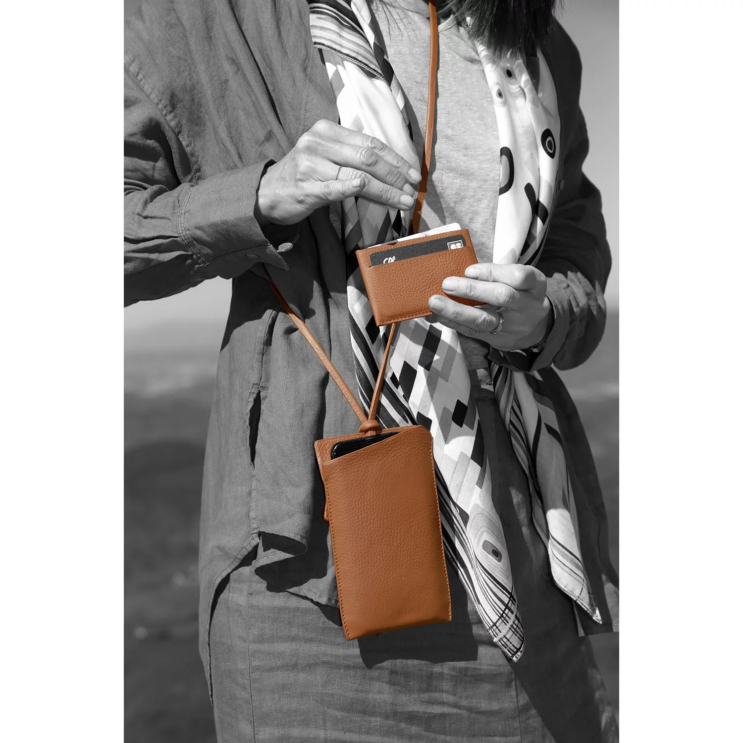 Une personne tient un portefeuille beige et une pochette pour téléphone, avec une carte partiellement visible. Elle porte une écharpe et une veste. L'arrière-plan est flou.