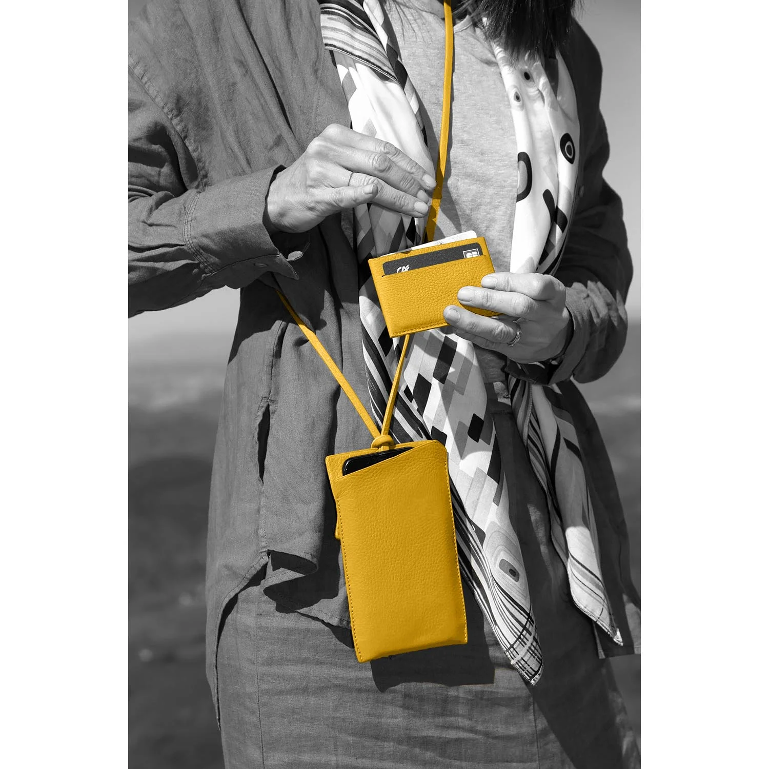 Personne tenant un portefeuille jaune et un étui assorti, portant une écharpe et un manteau à motifs dans un décor noir et blanc.