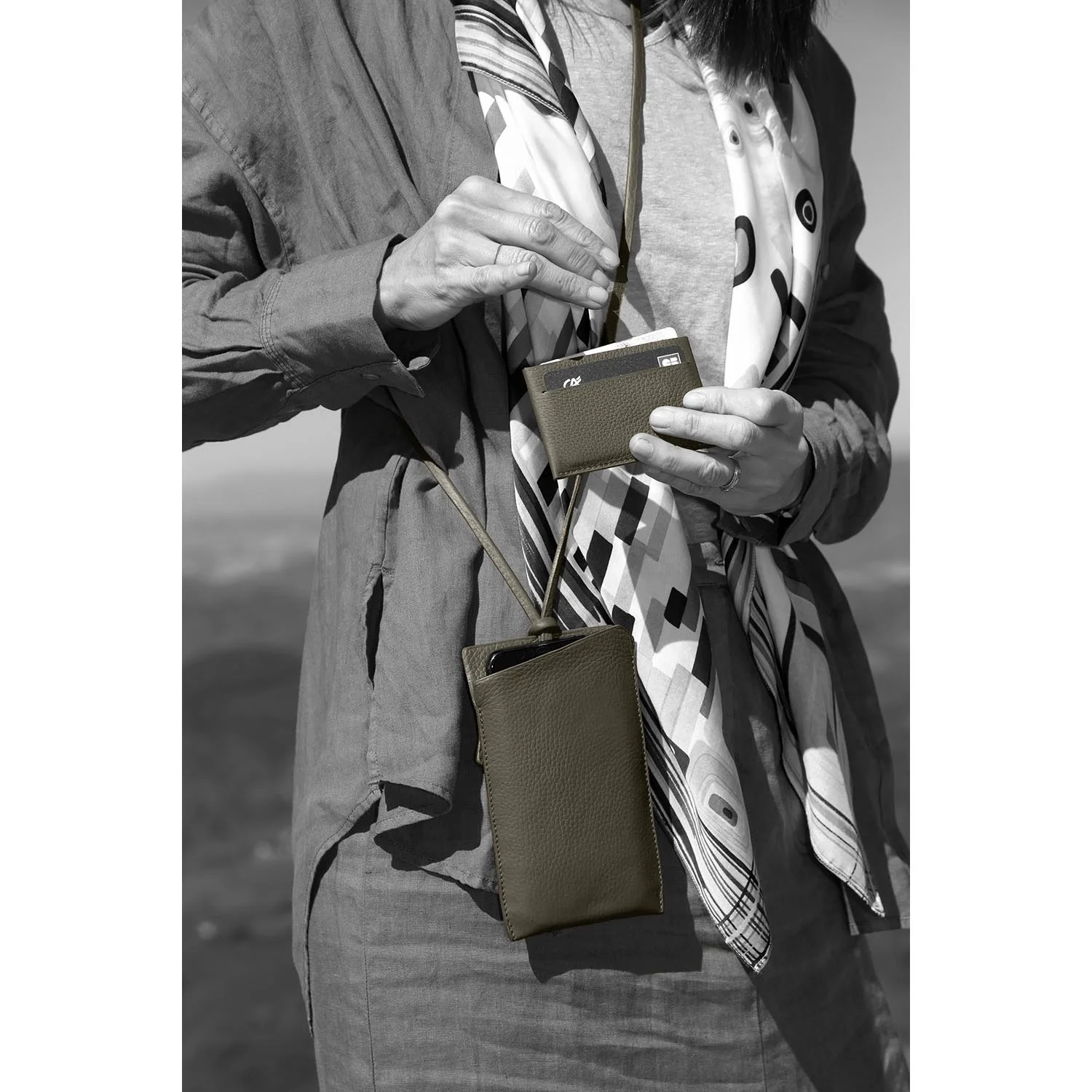 Personne tenant un petit portefeuille et un étui pour smartphone avec une sangle, portant une écharpe à motifs et une veste.