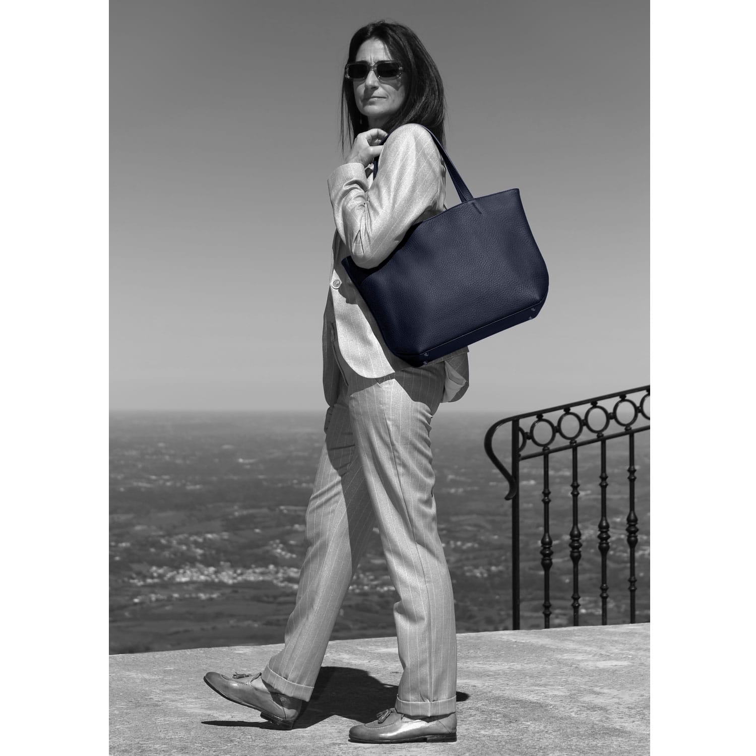 Une personne en costume et lunettes de soleil se tient à l'extérieur, tenant un grand sac, avec une vue panoramique en arrière-plan.