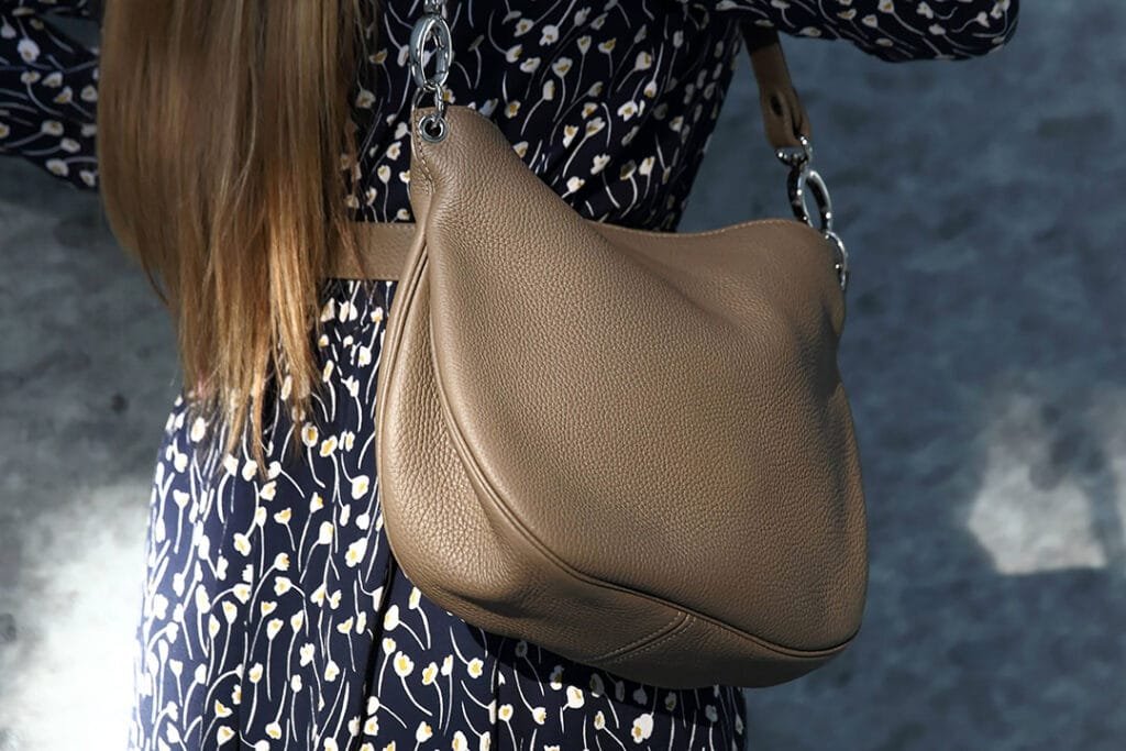 Une personne aux cheveux longs porte une robe à motifs et porte un sac à main en cuir beige sur son épaule.