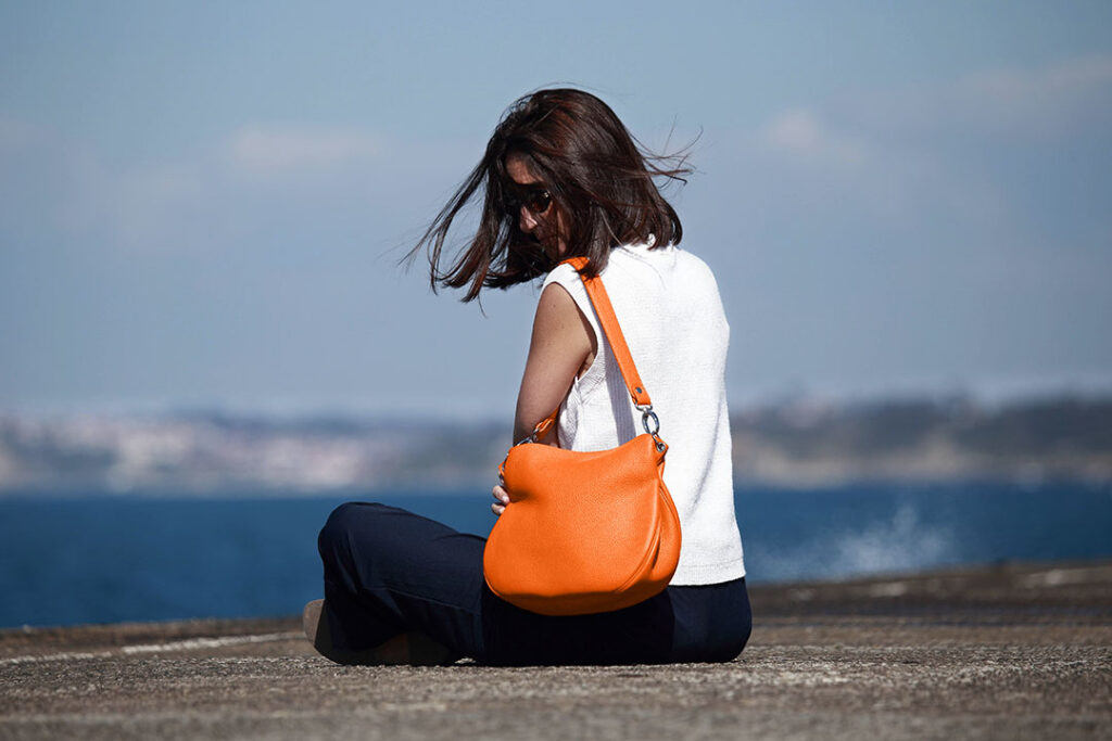 Une femme aux cheveux bruns mi-longs est assise sur une surface en béton près de l'eau. Elle porte des lunettes de soleil, un haut blanc, un pantalon sombre et un sac à main orange.