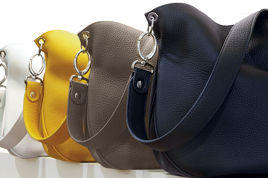 Quatre sacs à main en cuir blanc, jaune, taupe et noir, chacun doté d'une large lanière et d'un anneau argenté, sont alignés sur une surface réfléchissante.