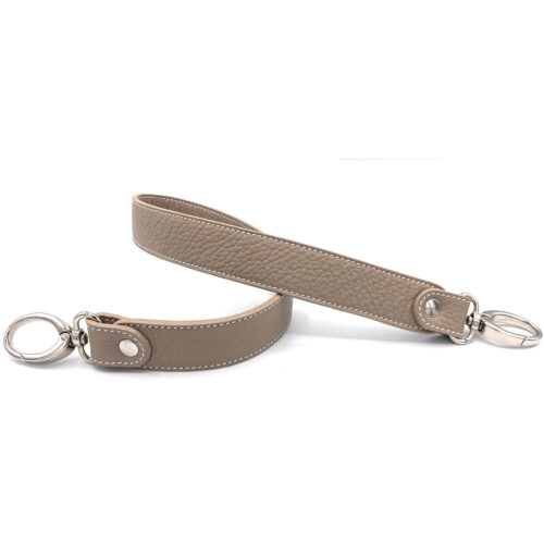 Sangle de sac en cuir taupe avec des coutures blanches, dotée de fermoirs métalliques argentés aux deux extrémités, présentée sur un fond blanc.