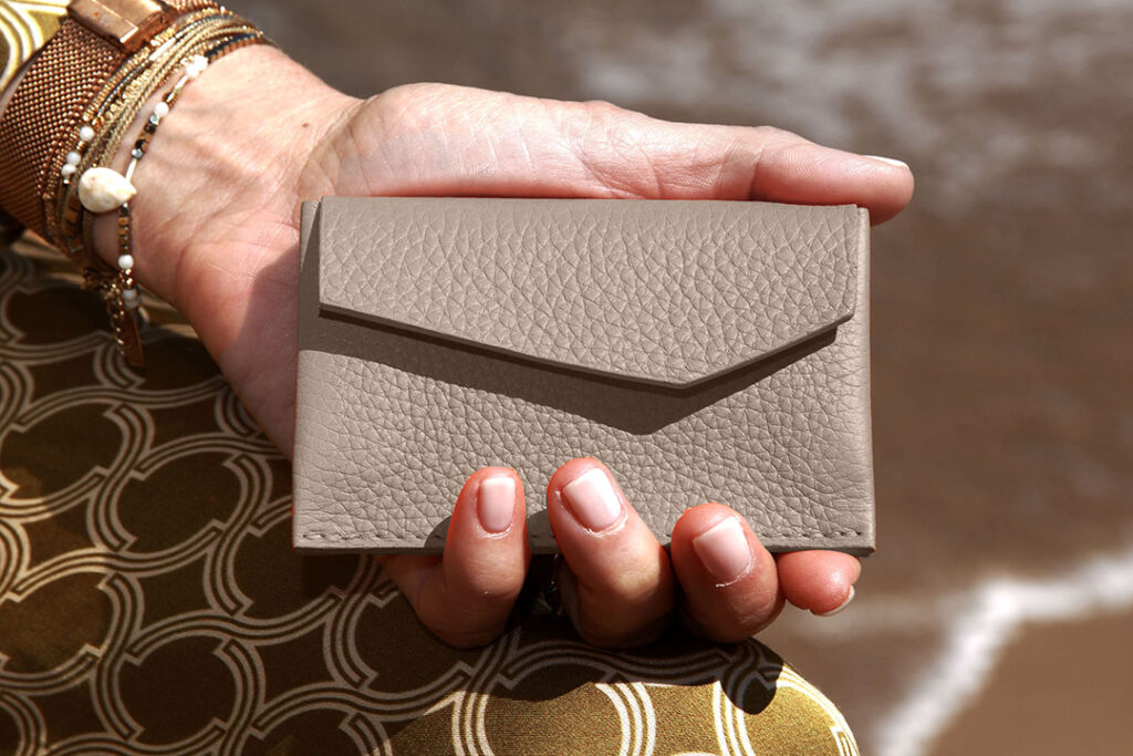 Une main tient un petit portefeuille en cuir beige texturé à l'extérieur, avec en arrière-plan un tissu à motifs, des bracelets et de l'eau floue.
