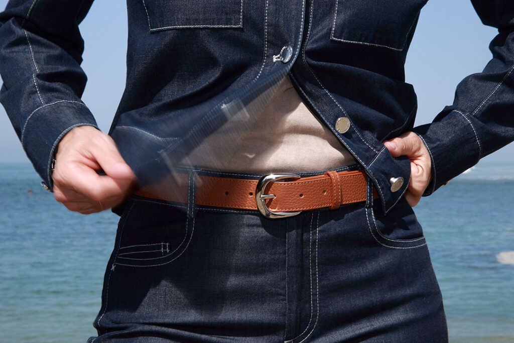Une personne vêtue d'un ensemble en jean et d'une ceinture en cuir grainé marron cognac détache rapidement sa ceinture près d'un plan d'eau.