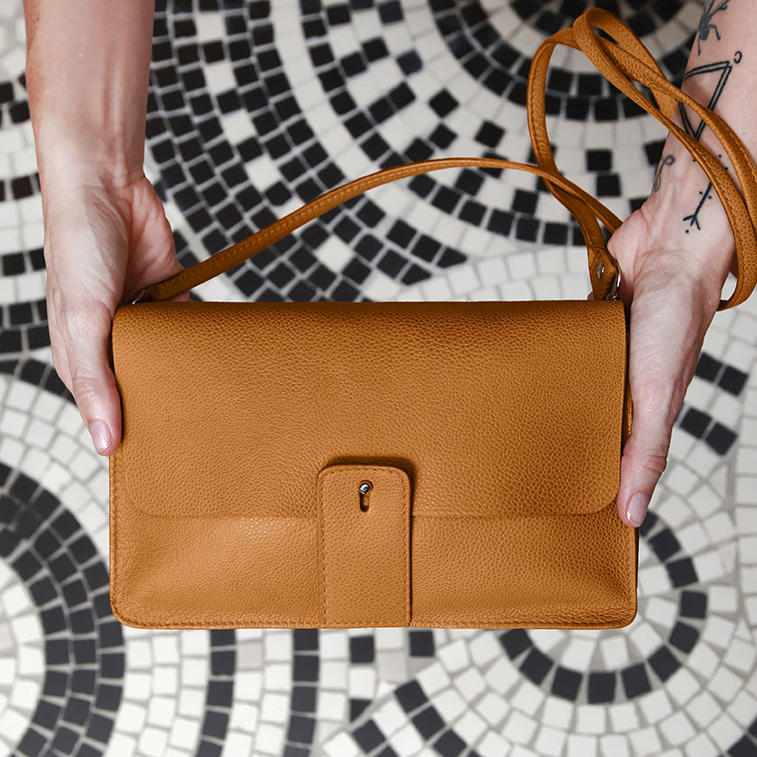 Des mains tenant un sac à bandoulière rectangulaire de couleur cognac, muni d'une fermeture à languette centrale.